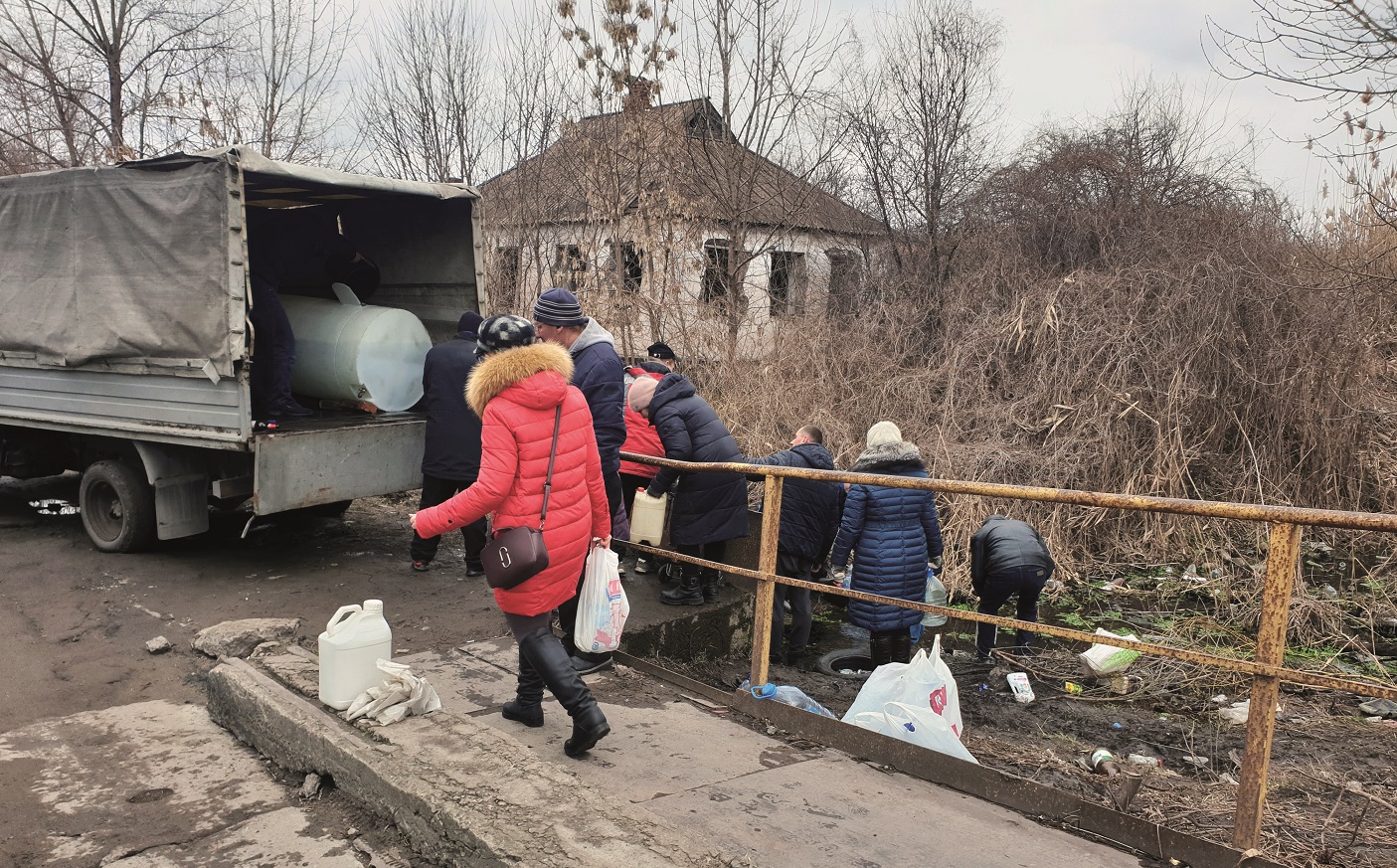 En hop av m&auml;nniskor verkar k&ouml;a till en lastbil med &ouml;ppet flak. En person i r&ouml;d l&aring;ng jacka passerar p&aring; v&auml;gen med en matkasse i handen.