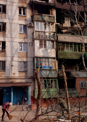 Tv&aring; m&auml;nniskor sopar g&aring;rden framf&ouml;r ett s&ouml;nderbombat hus. Skr&auml;p p&aring; marken, s&ouml;nderslitna tr&auml;d.