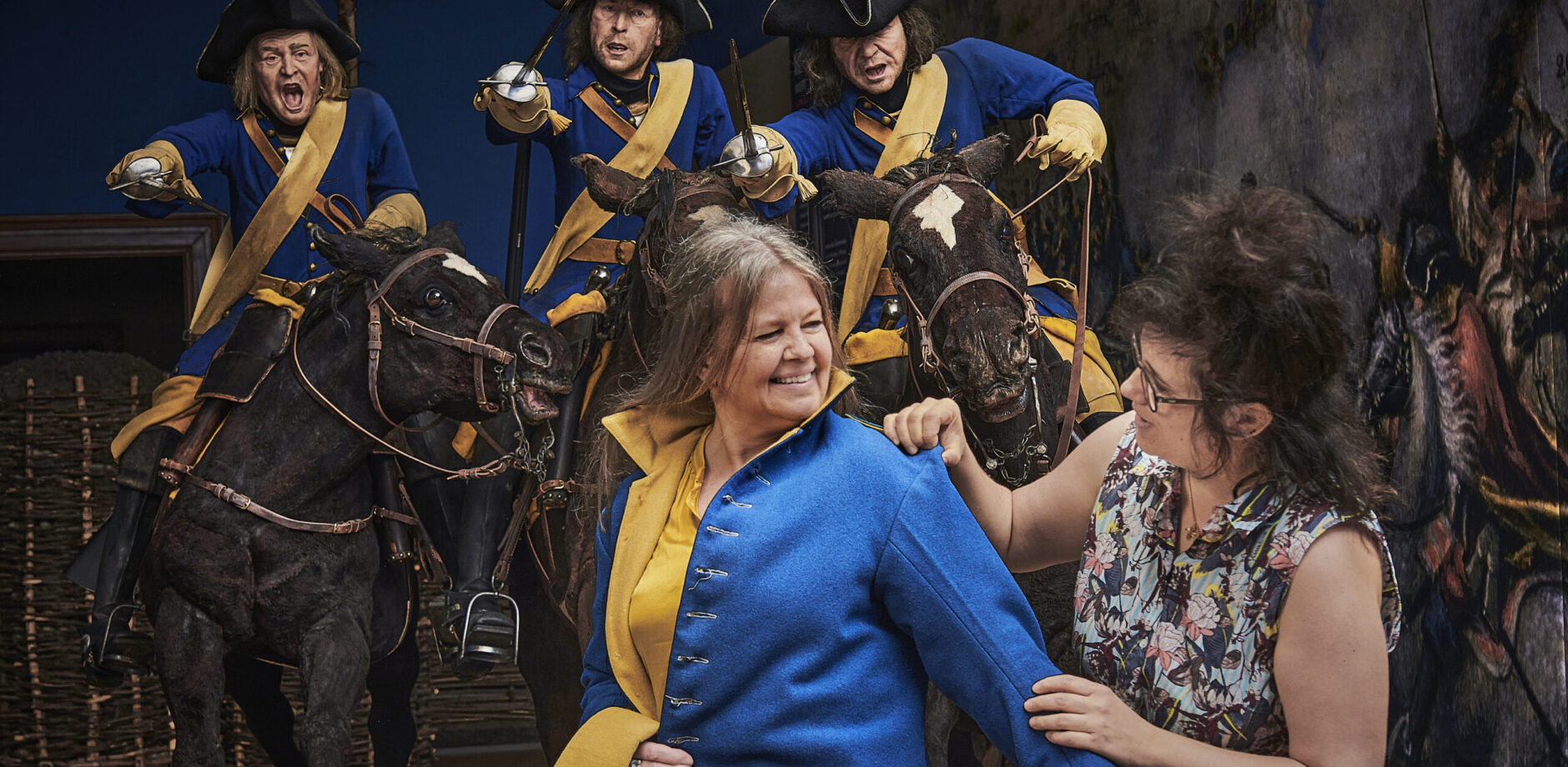  Två besökare testar Karolineruniform, bakom dem tre dockor till häst iklädda samma uniform.